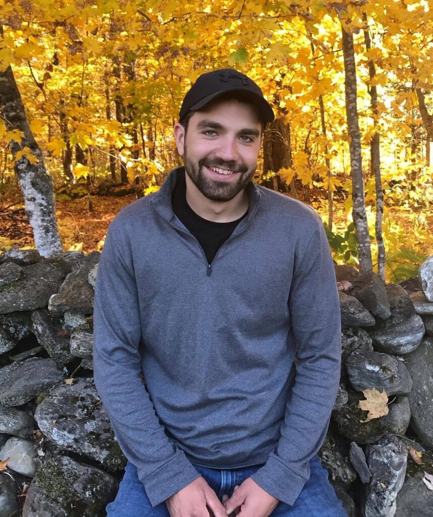Portrait of company owner Jake Atkinson sitting on stone wall in front of autumn birches for jake's mission page