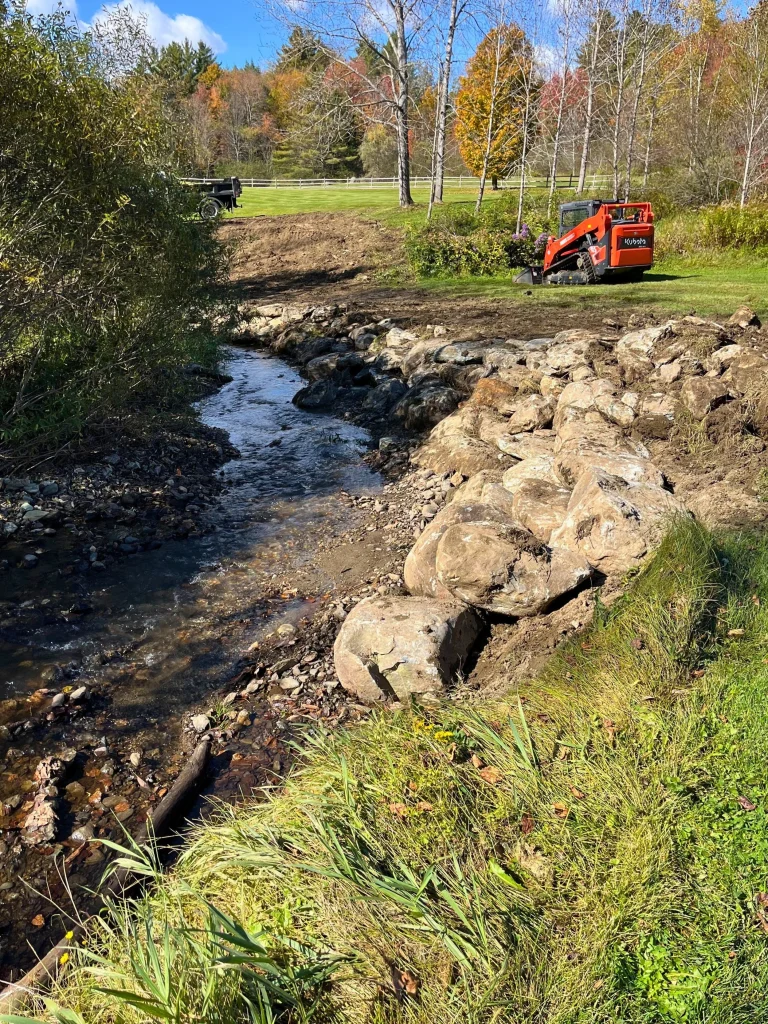 A small creek flowing through a landscaped yard with rocks retaining the edge, a Kubota compact loader on the grass, and autumn trees in the background.