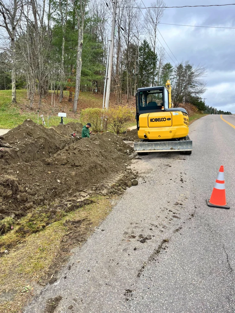 Construction worker operating a small yellow Kobelco excavator on the side of a rural road with a traffic cone nearby, digging a trench or hole next to the pavement.