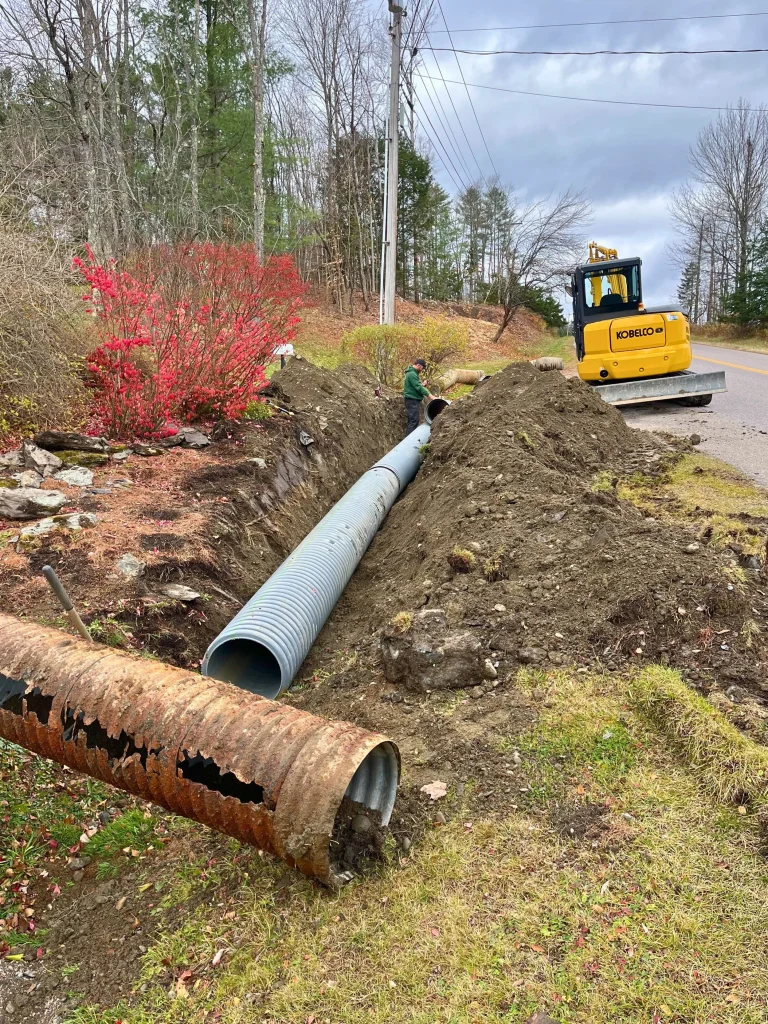Workers installing large gray utility pipe in an open trench along a roadside, with a yellow compact excavator nearby, under cloudy skies.